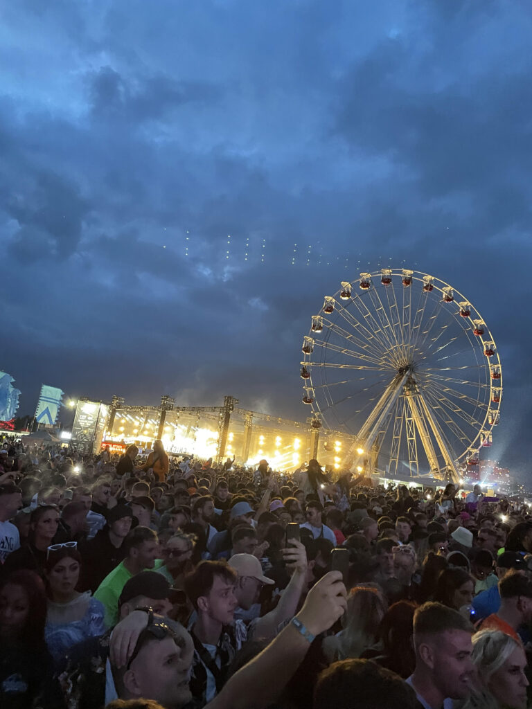 An image taken of a crowd at a festival with a stage and ferris wheel in the background.