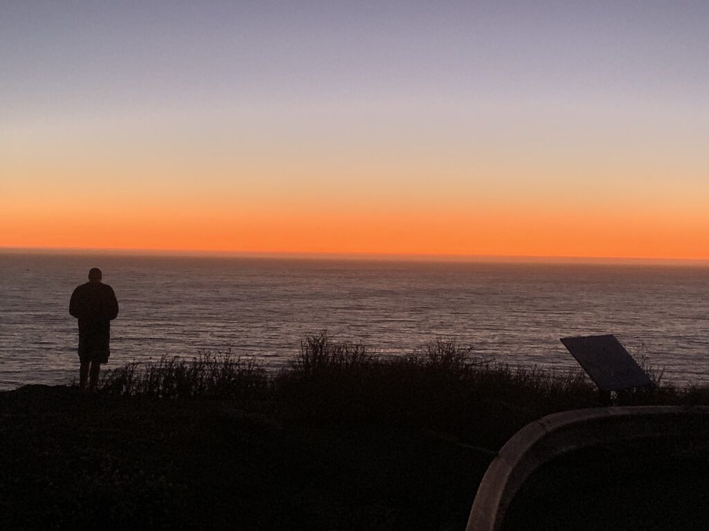 A man standing in the foreground of the ocean and sunset. 