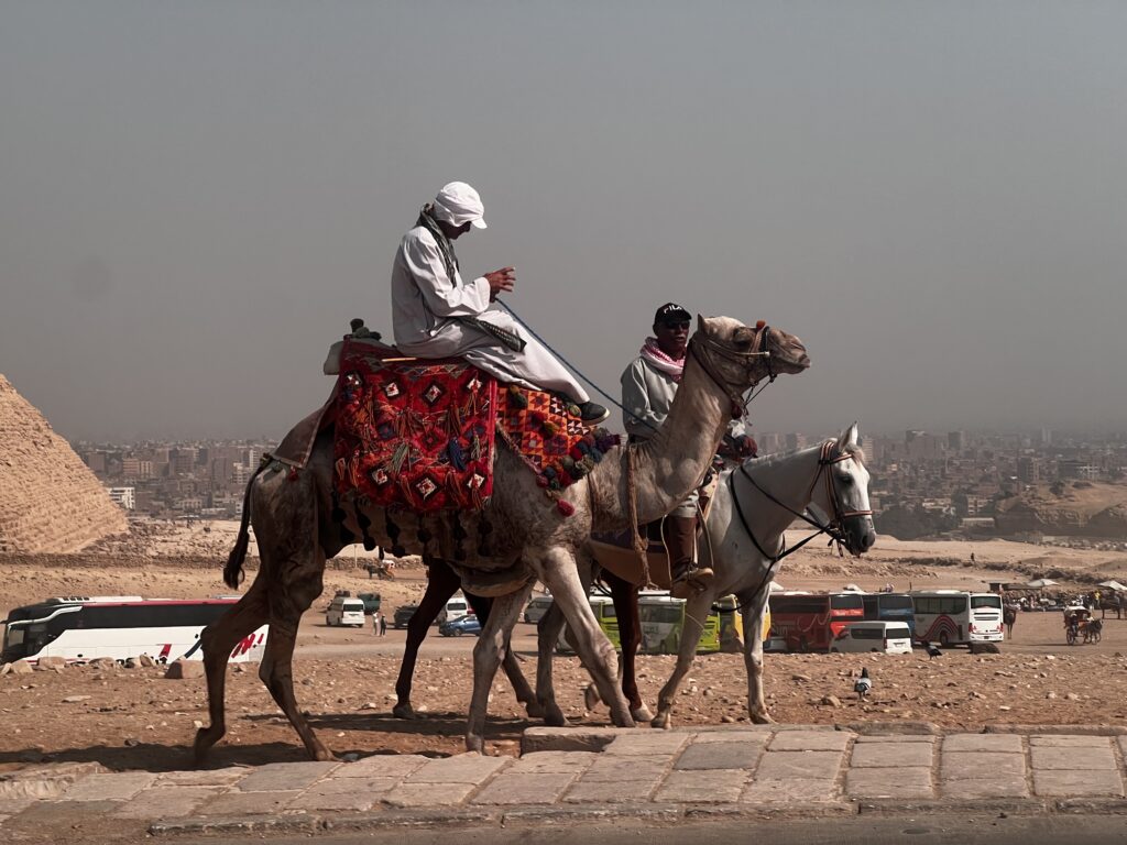 Two Egyptian men on a camel and a horse with the bottom of one of the Pyramids in the background.