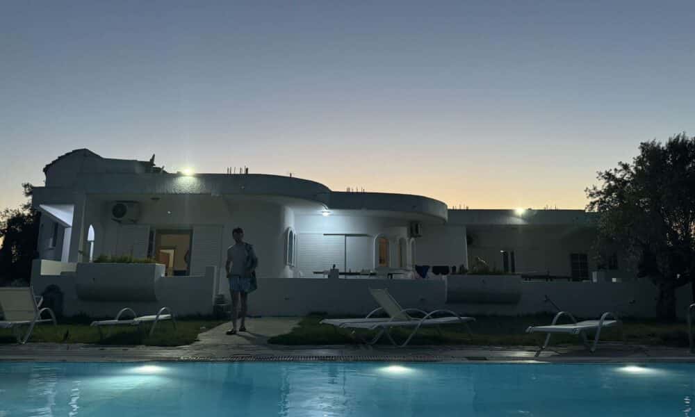 A landscape image of a Greek villa with a swimming pool in the foreground and a friend of the author strolling toward the pool.