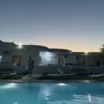 A landscape image of a Greek villa with a swimming pool in the foreground and a friend of the author strolling toward the pool.