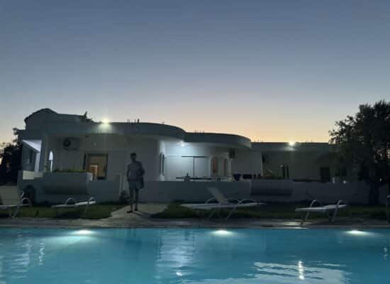 A landscape image of a Greek villa with a swimming pool in the foreground and a friend of the author strolling toward the pool.