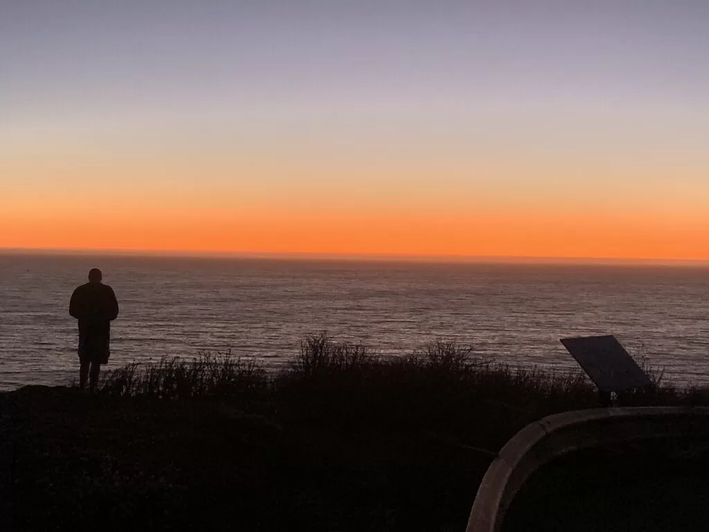 A man standing in the foreground of the ocean and sunset. 