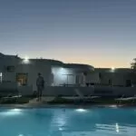 A landscape image of a Greek villa with a swimming pool in the foreground and a friend of the author strolling toward the pool.